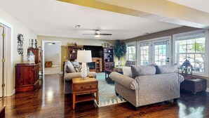 A spacious living room with hardwood floors, a ceiling fan, and large windows. - Private Pool & Hot Tub at Luxe Ringgold Retreat (Ringgold)