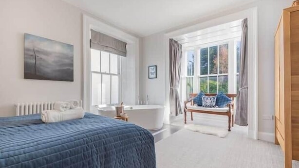 A bedroom with a large bed, a bathtub, and a window with curtains. - Peregrine Apartment Bowness (Bowness-on-Windermere)