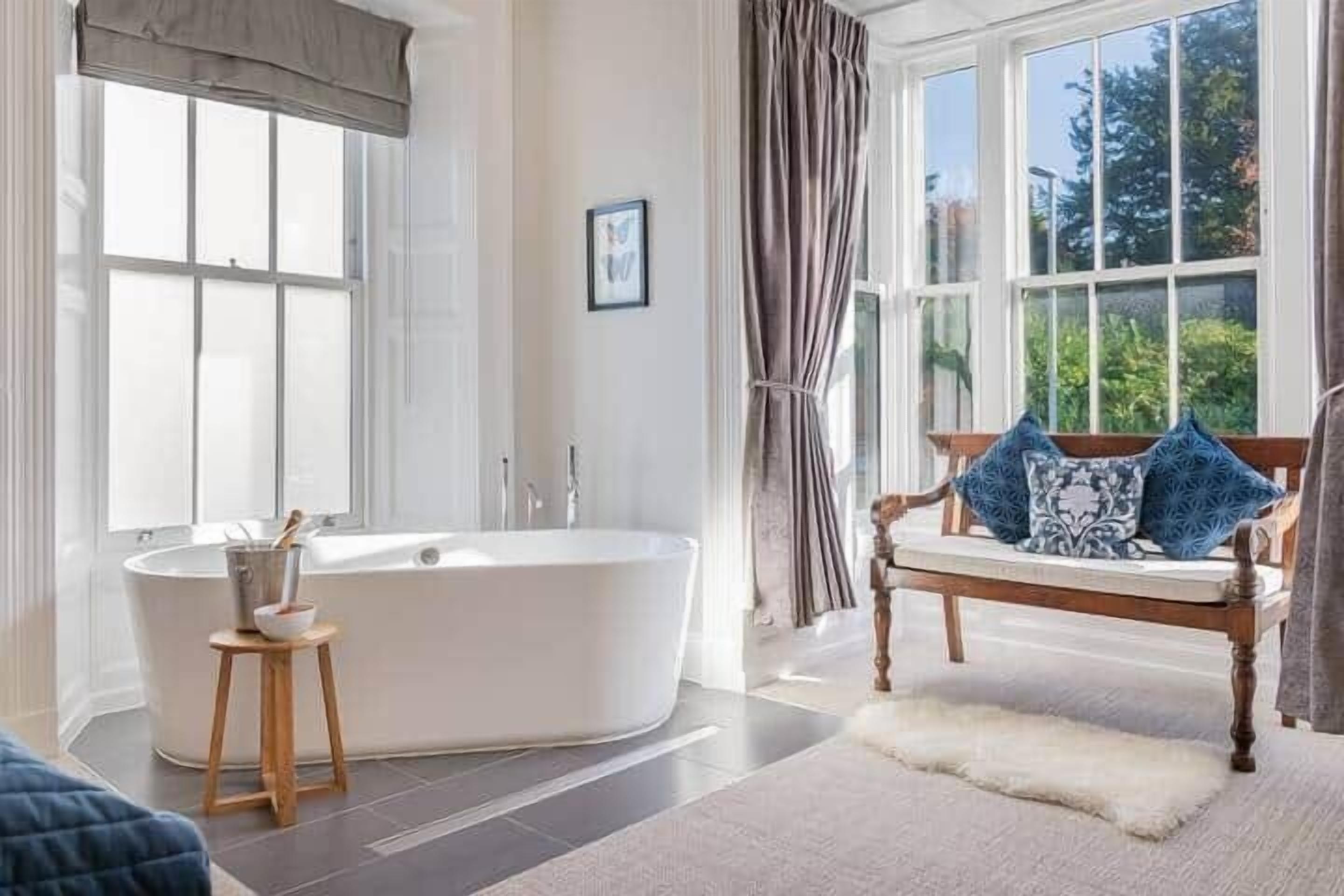 A modern bathroom with a freestanding bathtub, a wooden stool, and a bench with decorative pillows.