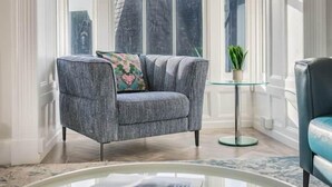 A grey armchair with a floral pillow, a glass coffee table with a book, and a small potted plant on a glass table. - Peregrine Apartment Bowness (Bowness-on-Windermere)