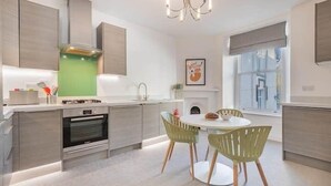 A modern kitchen with a built-in oven, a dining area with two chairs, and a large window providing natural light. - Peregrine Apartment Bowness (Bowness-on-Windermere)