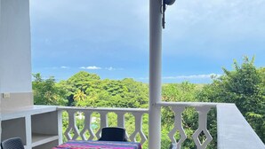 A covered outdoor seating area with a table and chairs, a white railing, and a view of lush greenery and the sea.