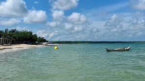 A calm beach with clear turquoise water, a small boat, and a distant shoreline with vegetation.