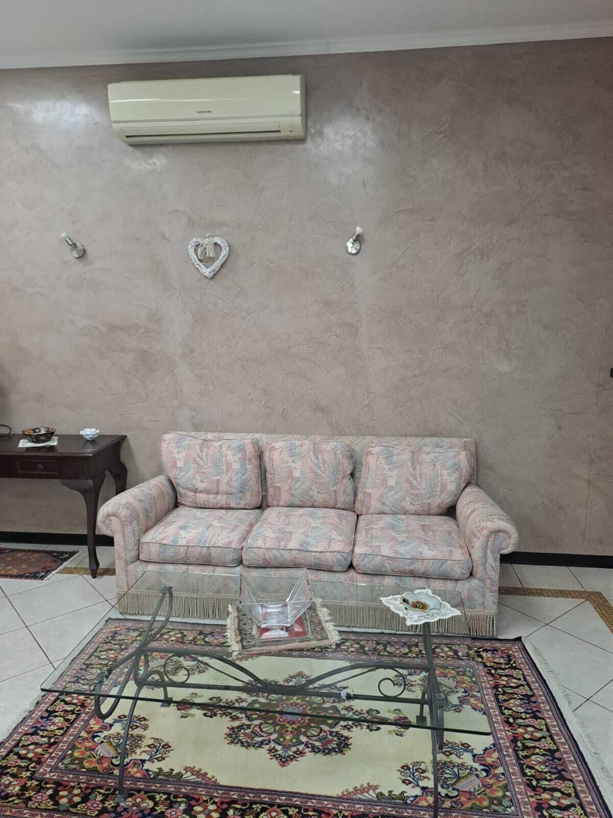 A living room with a floral-patterned sofa, a glass coffee table, a patterned rug, and an air conditioning unit on the wall.