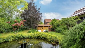Une maison avec un étang, entourée d’une végétation luxuriante et d’arbres.