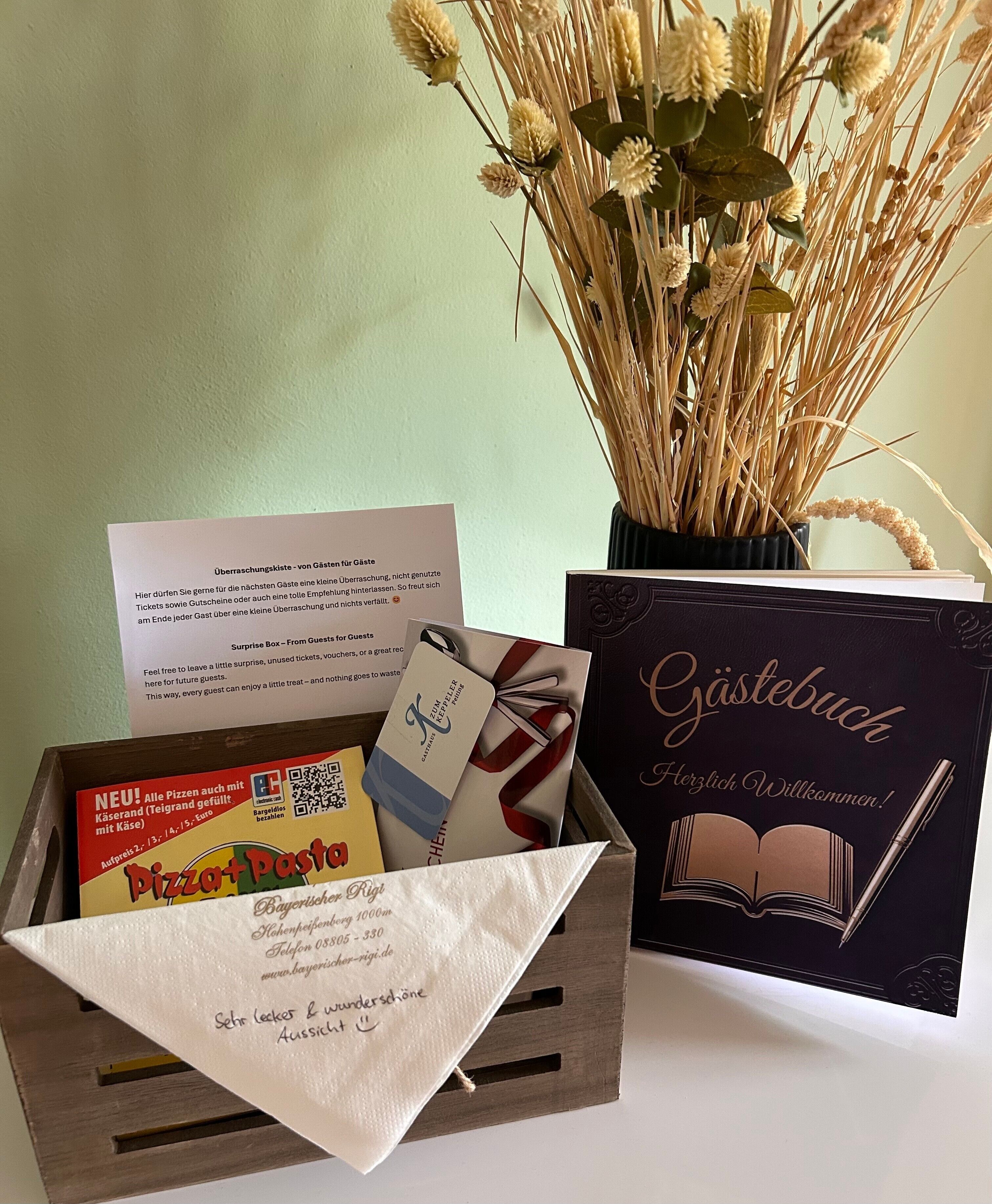 A wooden box with a pizza box, a gift card, a card with a book illustration, and a vase with dried plants.
