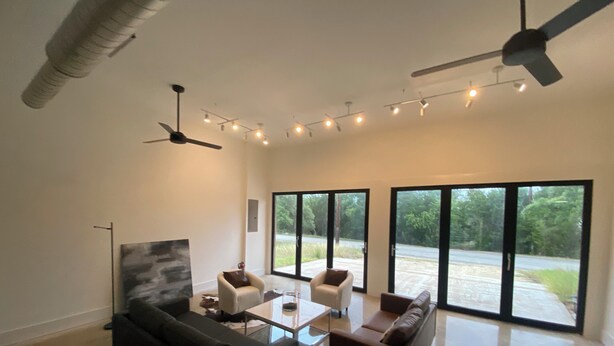 Un salon spacieux avec un canapé noir, deux fauteuils, une table basse et un ventilateur de plafond. De grandes fenêtres offrent une vue sur l’extérieur.