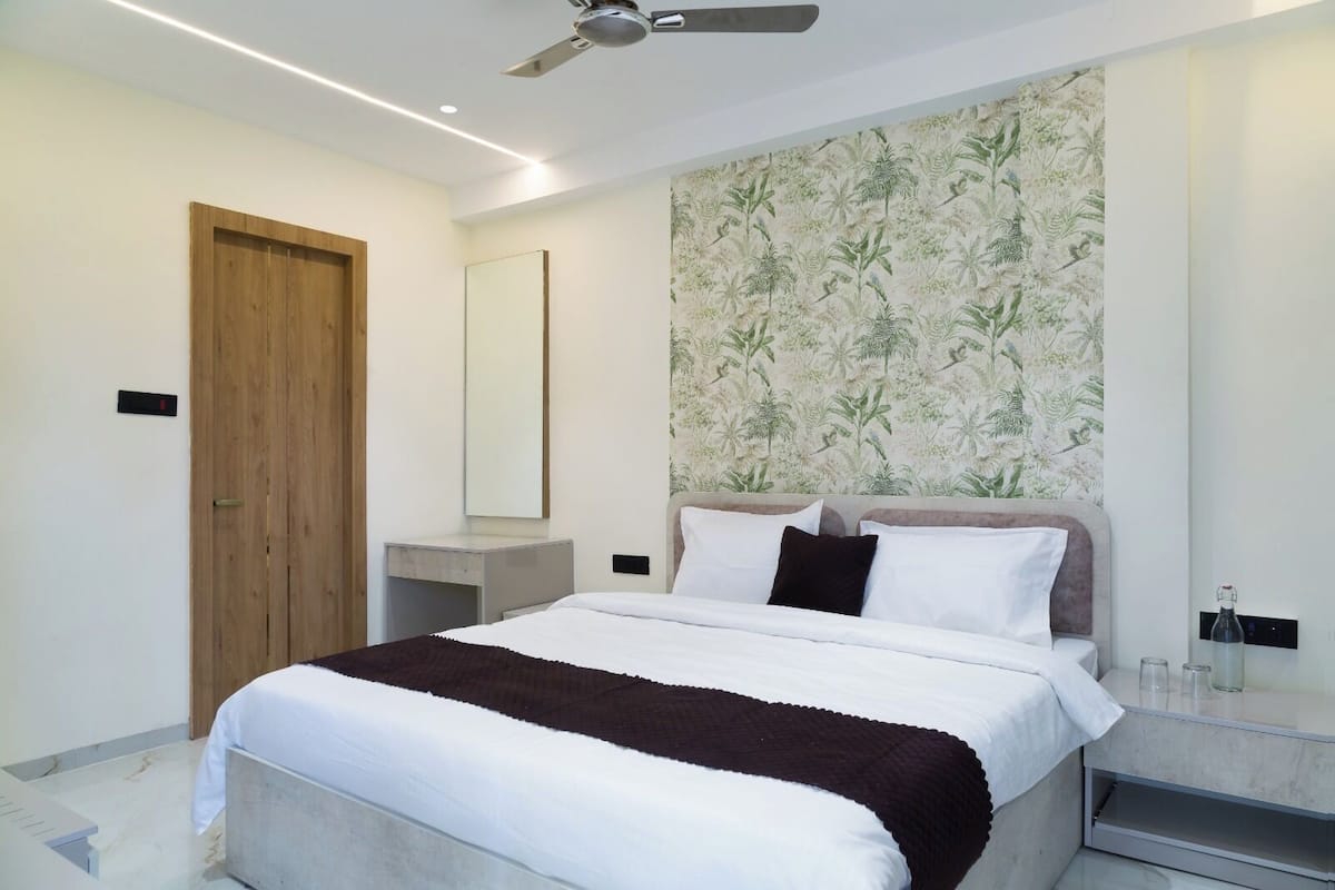 A modern bedroom with a bed, a wooden door, a desk, a mirror, and a wall with a tropical wallpaper.