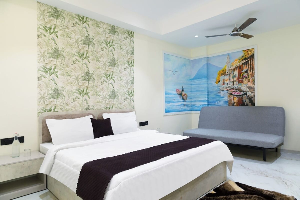 A bedroom with a bed, a gray sofa, a ceiling fan, and a large painting of a cityscape.