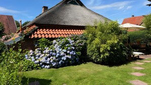 Una casa con techo de paja, chimenea de ladrillo y un jardín con hortensias y bambú.