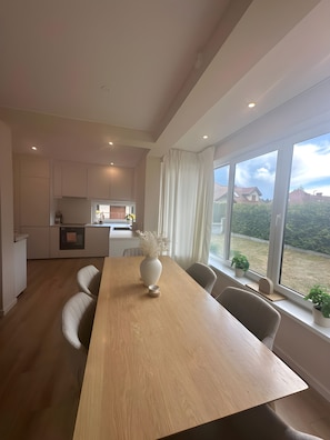 A modern kitchen with white cabinets, a wooden dining table, and a view of the garden through large windows.