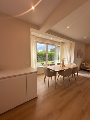 A modern kitchen with white cabinets, a dining area with a wooden table and chairs, and large windows with curtains.