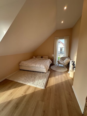 A bedroom with a sloped ceiling, a large bed, a chair, a small table, and a view of a building outside.