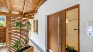 Une porte en bois avec une plaque portant un numéro, une structure en bois à lattes ornée de plantes, et un pot de fleurs posé sur le sol.