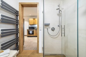A modern bathroom with a glass shower enclosure, a wooden bench, and a visible kitchen area in the background.