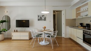 Una cucina moderna con mobili bianchi, un'area pranzo con un tavolo rotondo e sedie, un televisore a schermo piatto e una pianta in vaso.