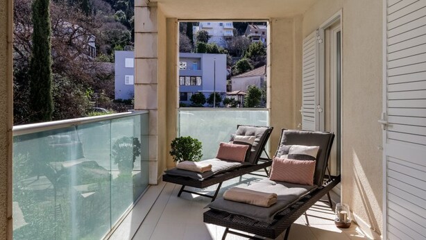 Un balcone con due poltrone, ringhiera in vetro e vista su edifici e alberi.