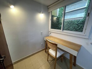Une petite pièce avec un bureau et une chaise, une fenêtre donnant sur un espace verdoyant et un sol carrelé.