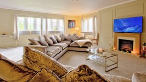 A living room with a large sectional sofa, a flat-screen TV, a fireplace, and large windows. - River View Cottage (Skipton)