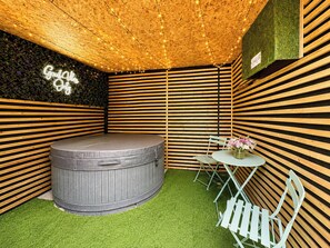 Una habitación con techo de listones de madera, una bañera de hidromasaje circular, una pequeña mesa redonda con un florero y un letrero de neón.