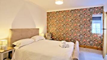 Un dormitorio con cama, mesitas de noche, lámpara y papel tapiz con motivos florales.