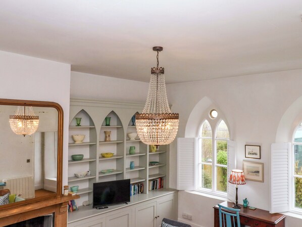 A room with a television on a white cabinet, a chandelier, shelves with decorative items, and a window with shutters.