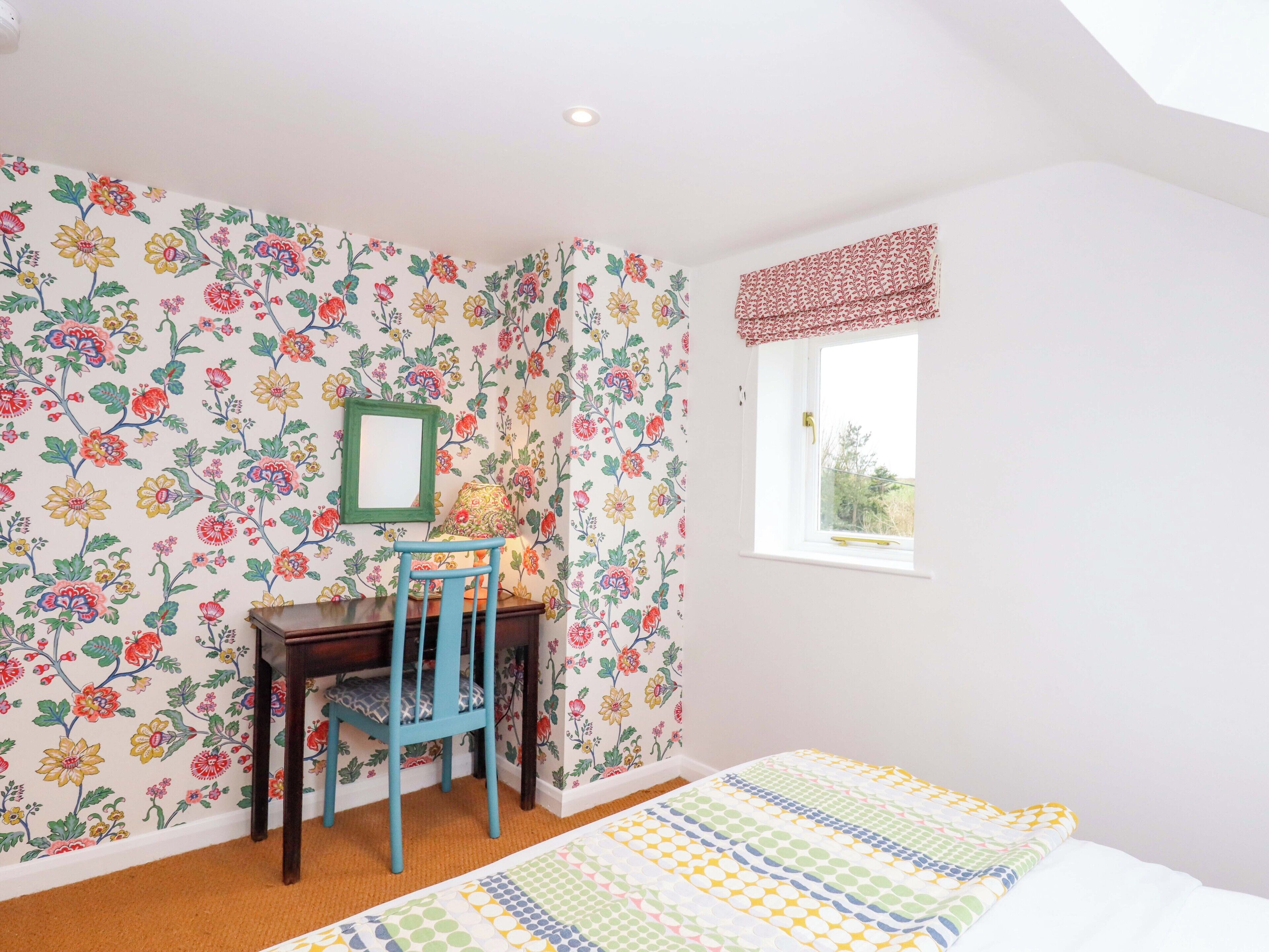 Een slaapkamer met bloemenbehang, een houten bureau met stoel, een raam met rode vouwgordijnen, en een bed met een bedovertrek met dessin.