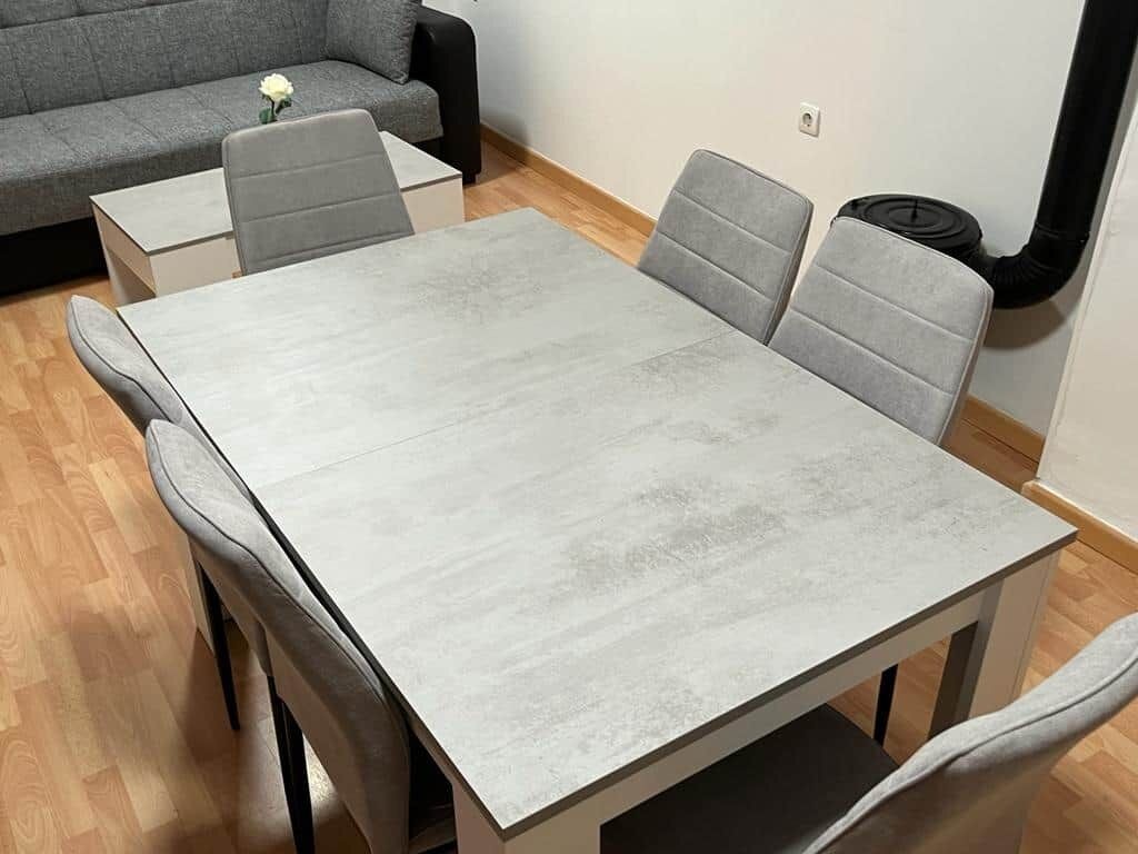 A dining area with a rectangular table and four grey upholstered chairs. A grey sofa is against the wall, and a small white table is visible in the background.