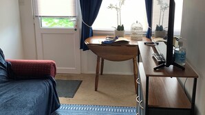 Una habitació amb escriptori, cadira i llit. Hi ha una finestra amb cortines, una planta i un vas amb flors.