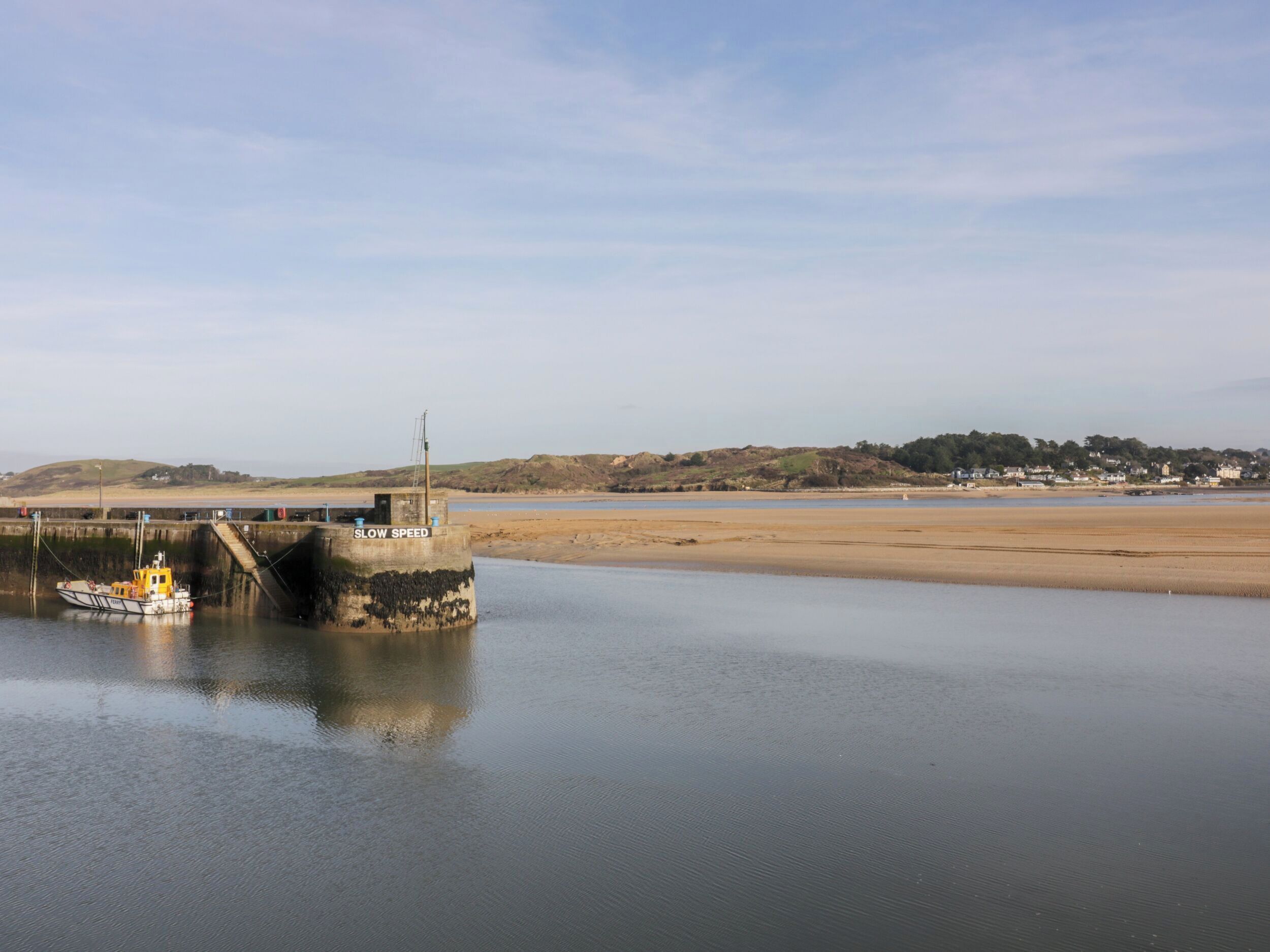 Un paysage côtier avec un bateau amarré à un quai portant l’inscription « ALLURE RÉDUITE », une plage de sable et une colline lointaine parsemée de bâtiments.