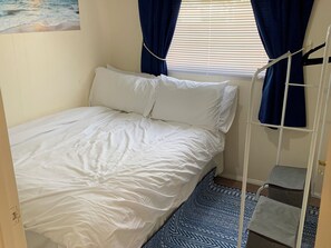 Una habitación pequeña con cama individual, cubrecama blanco, alfombra estampada en azul y ventana con cortinas azules.