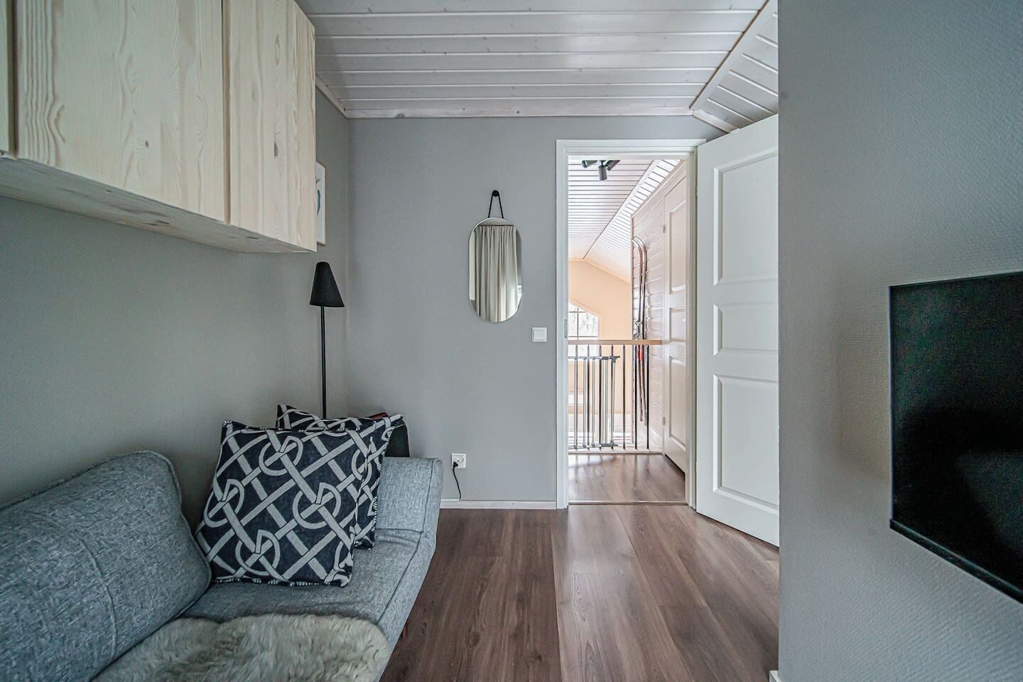 Ein modernes Wohnzimmer mit einem grauen Sofa, einem Schwarz-Weiß-Muster-Sitzkissen, einer Stehlampe und einem an der Wand montierten Fernseher.