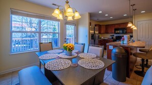 A dining area with a table set for six, a kitchen in the background, and a view of the outdoors through large windows.