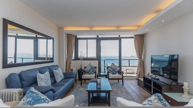 A modern living room with a sofa, chairs, a coffee table, a TV, and large windows offering a view of the sea. - Secure One Bed Uninterrupted Ocean Views (Cape Town)