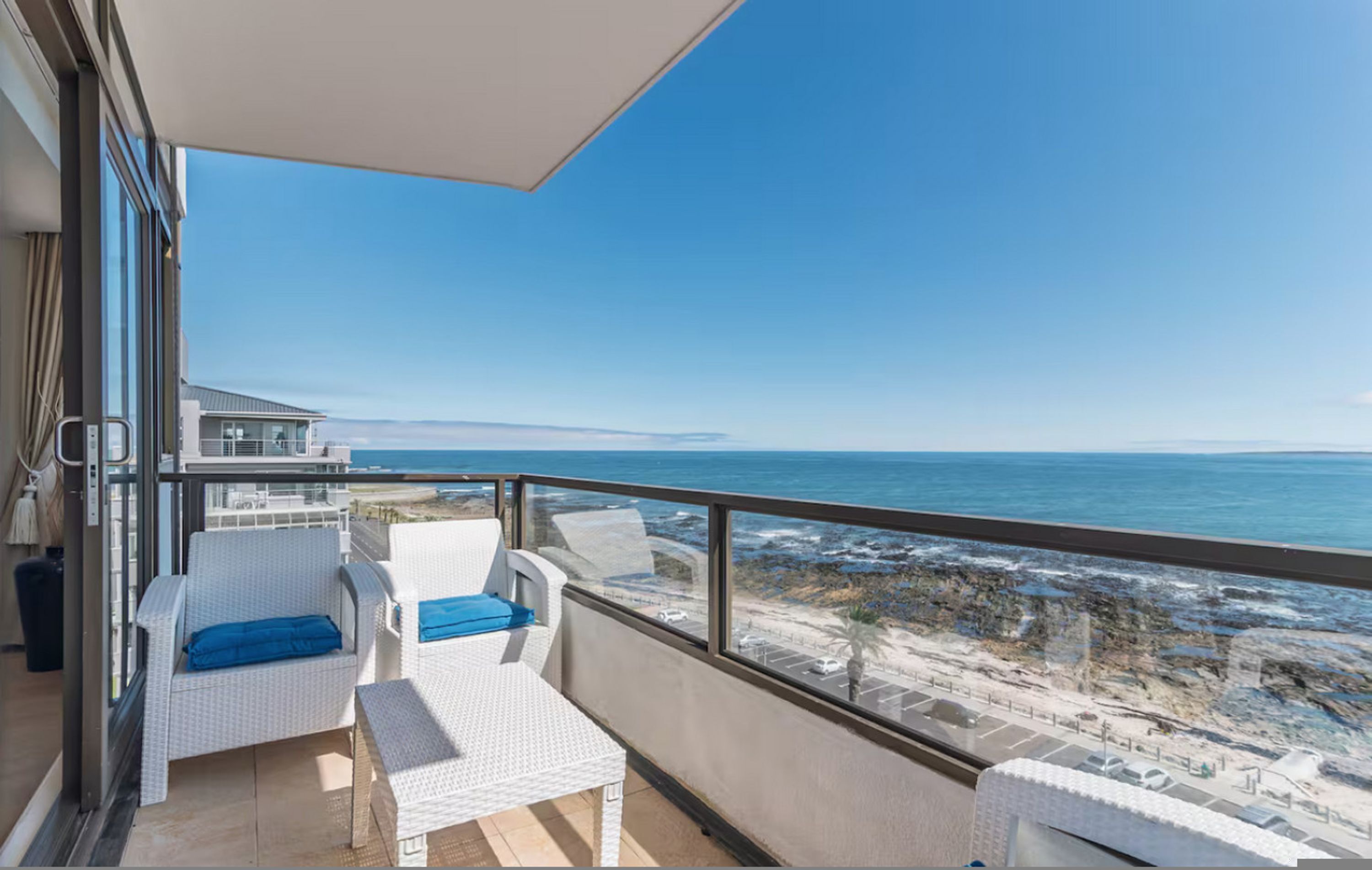 A balcony with white wicker furniture, a clear view of the ocean, and a rocky beach in the background.