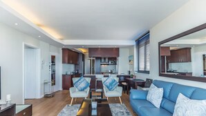 A modern living room with a blue sofa, two armchairs, a coffee table, and a kitchen in the background. - Secure One Bed Uninterrupted Ocean Views (Cape Town)