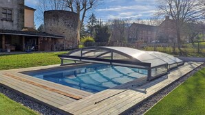 Ein Schwimmbecken mit Glasüberdachung, umgeben von einer Holzterrasse und einer Rasenfläche.