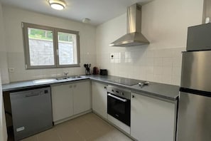 Eine kleine Küche mit weißen Schränken, einem Edelstahlkühlschrank, einem Geschirrspüler und einem Fenster für natürliches Licht.