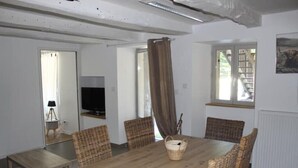 A dining area with a wooden table and wicker chairs, a television on a stand, and a window with a view of a staircase. - SIBO GITE apartment 4 pers, swimming pool, nature and city (Boulazac Isle Manoire)
