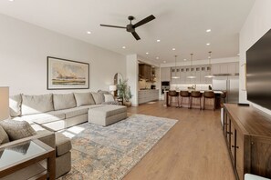 A modern living room with a sofa, coffee table, and a large rug. There is a dining area with bar stools and a kitchen with a refrigerator and cabinets. - Coral Rock Retreat 7 Bedroom Home (St. George)