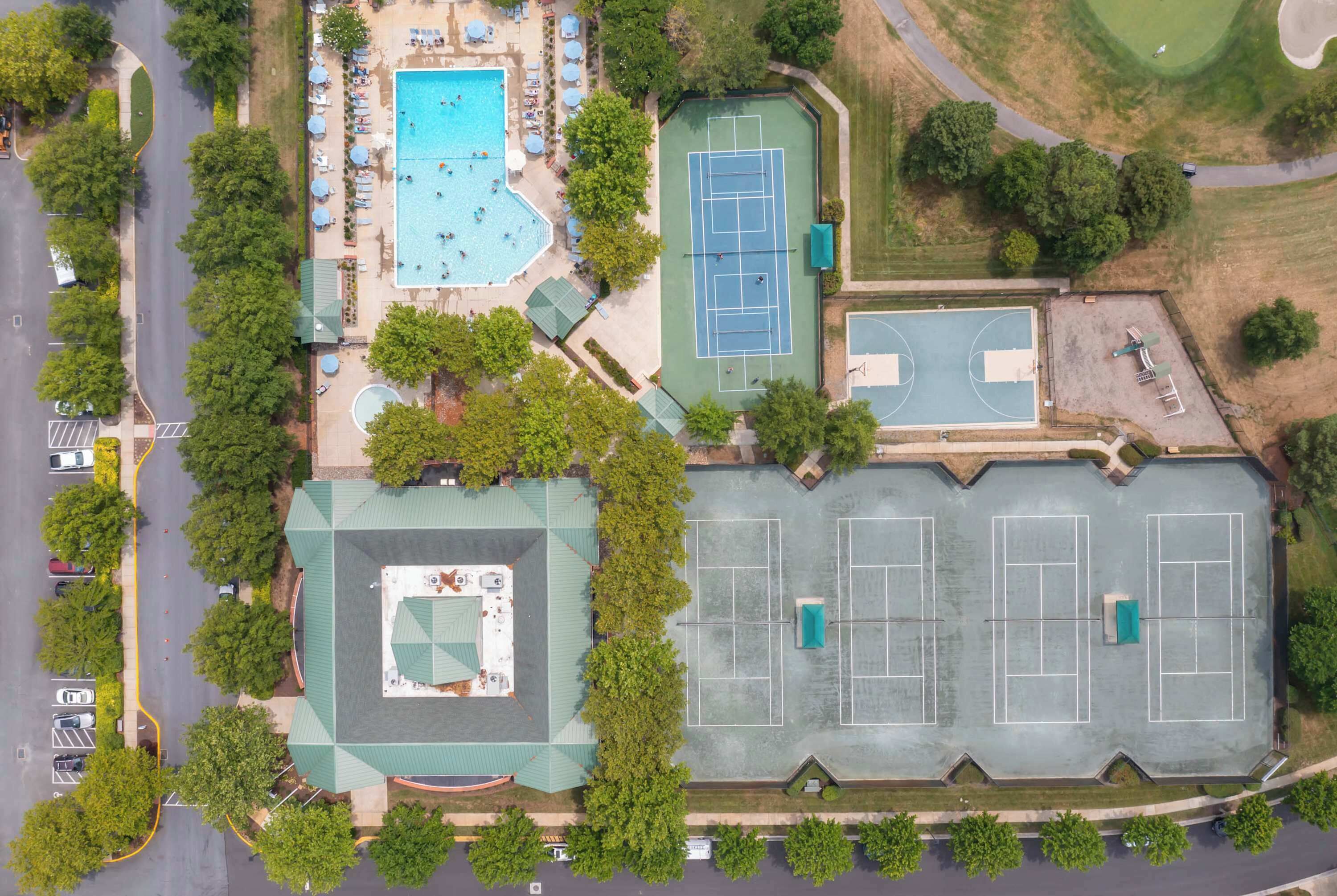 Luchtbeeld van een complex met een zwembad, tennisbanen en een parkeerplaats.