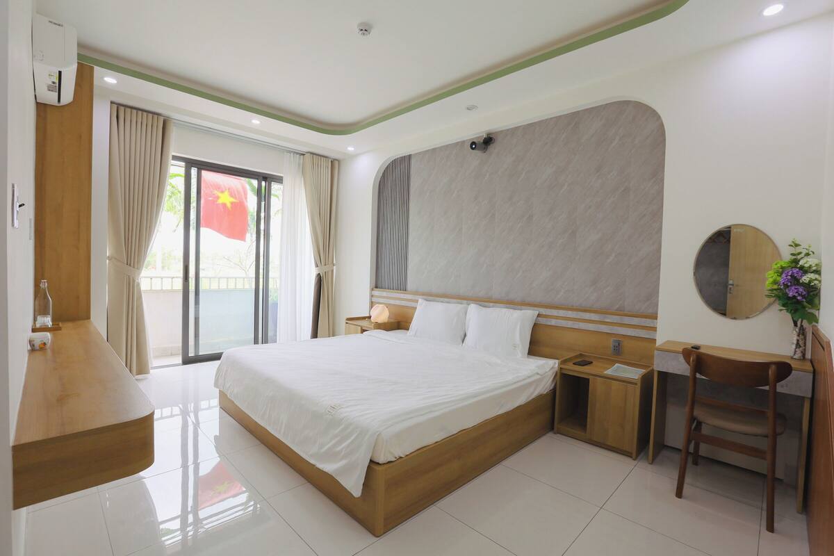 A hotel room with a large bed, wooden headboard, bedside tables, a chair, and a view of the outdoors.
