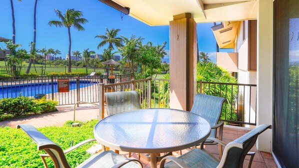 A balcony with a glass table and chairs overlooking a swimming pool, surrounded by palm trees and a fence. - Ground-Floor Maui Eldorado 1 Bedroom Condo Steps to Pool & BBQ | Unit C103 (Kaanapali)