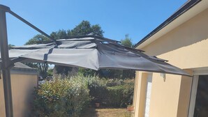Een terras buiten met een tafel en stoelen, een grote parasol en uitzicht op de achtertuin.