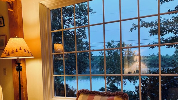 A cozy room with a window seat by a large window overlooking a body of water. A lamp and a book are placed on the seat. - Classic Maine Waterfront Home on the Back River – Trevett (Boothbay)