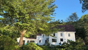 A two-story white house with a dark roof, surrounded by lush greenery and trees. - Classic Maine Waterfront Home on the Back River – Trevett (Boothbay)
