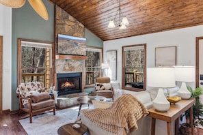 A cozy living room with a fireplace, comfortable seating, and large windows offering a view of the outdoors.