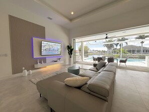 A modern living room with a large flat-screen TV mounted on the wall, a comfortable sofa, a dining area with chairs, and a view of a swimming pool and outdoor area. - Villa Elements - new, on water, pool, spa, modern (Cape Coral)