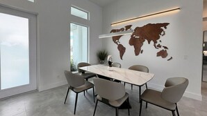 A modern dining area with a large marble table and six chairs, a world map on the wall, and a large window providing natural light. - Villa Elements - new, on water, pool, spa, modern (Cape Coral)