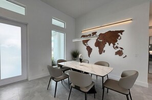 A modern dining area with a large marble table and six chairs, a world map on the wall, and a large window providing natural light. - Villa Elements - new, on water, pool, spa, modern (Cape Coral)